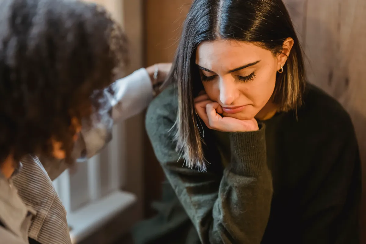 A friend placing a comforting hand on the shoulder of a pensive young woman